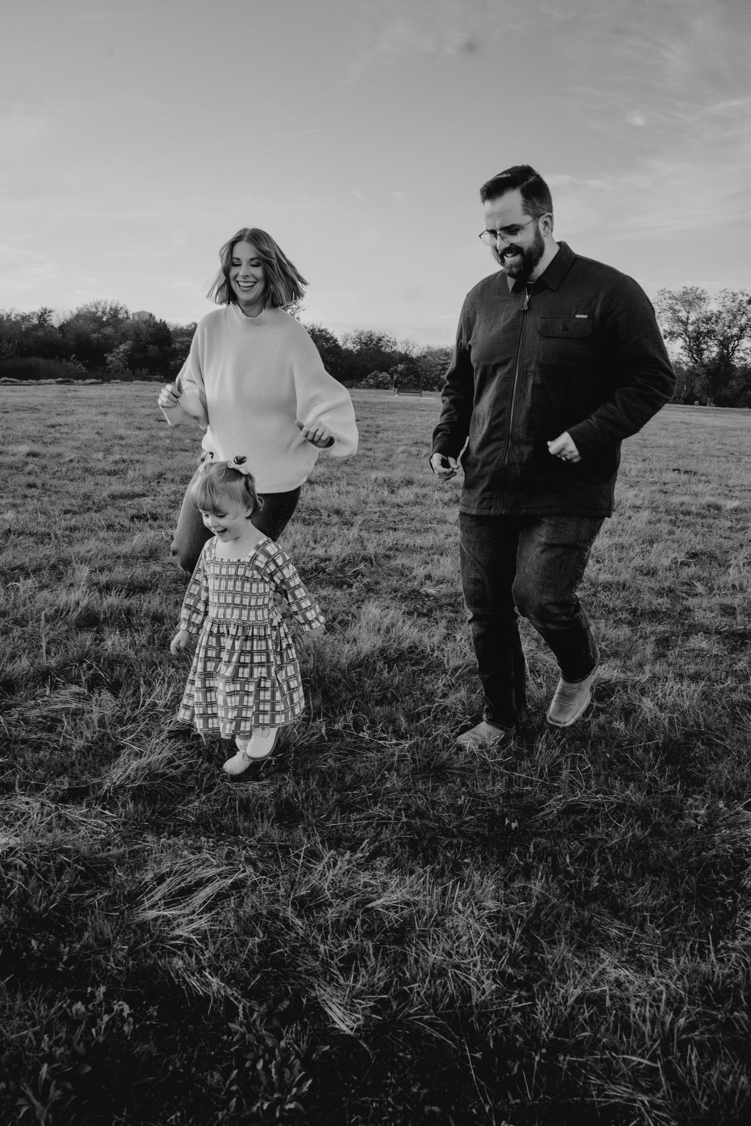 Black and white photo of happy family with toddler running together in an open field at dusk.