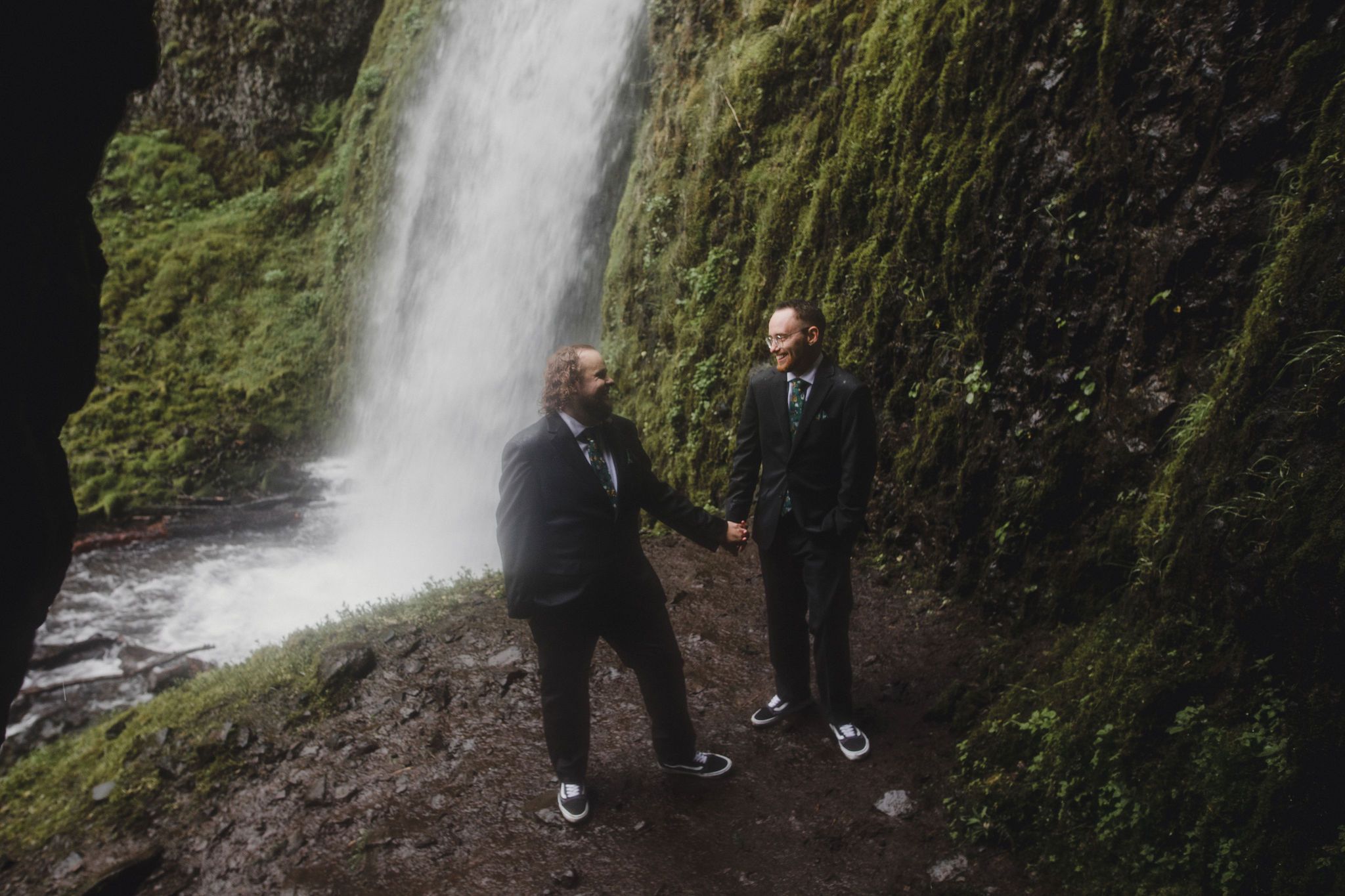Two grooms hold hands in front of a stunning waterfall surrounded by lush green moss during their outdoor elopement.