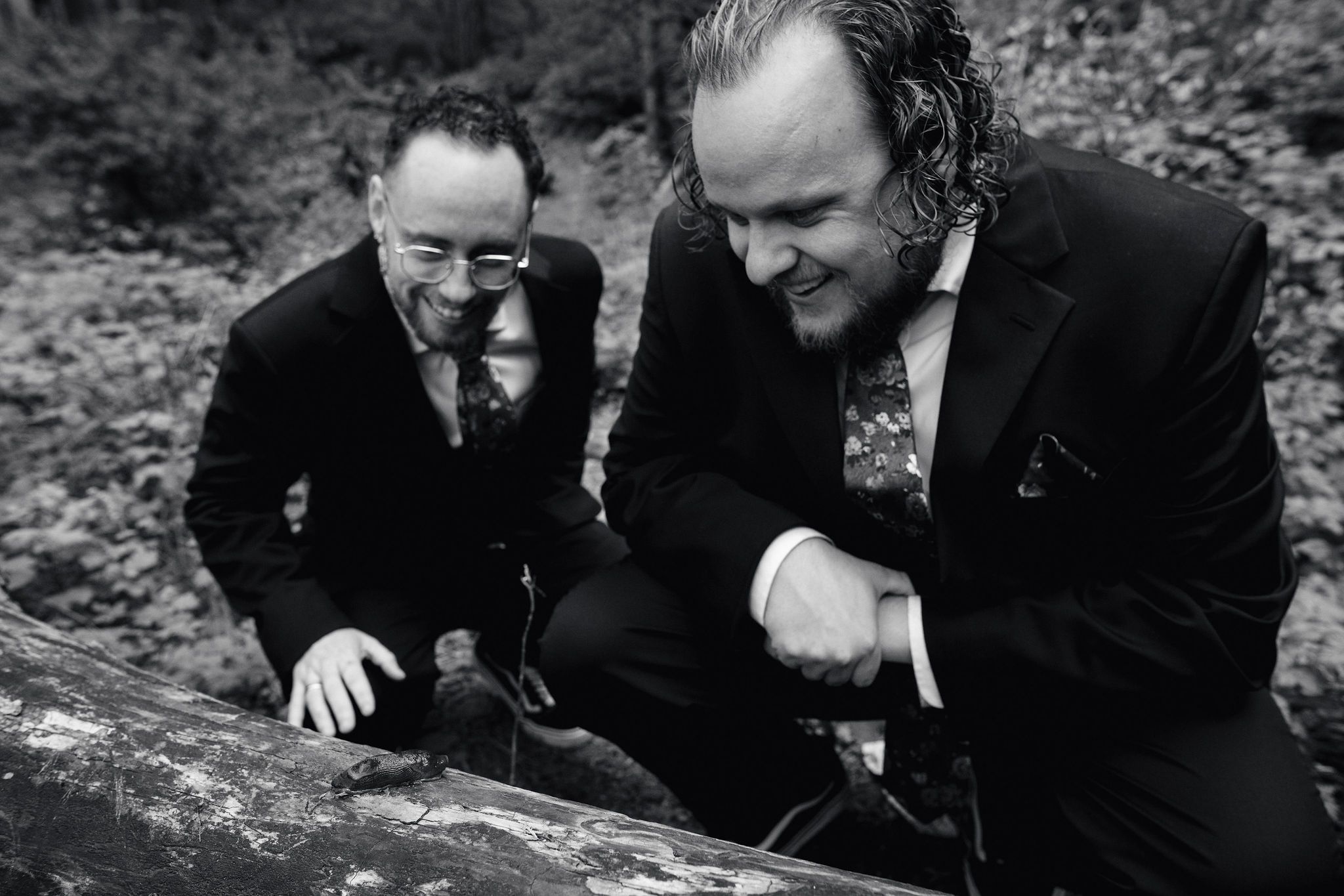 Two men in dark suits crouch outdoors in a black and white photo among rocks and trees.