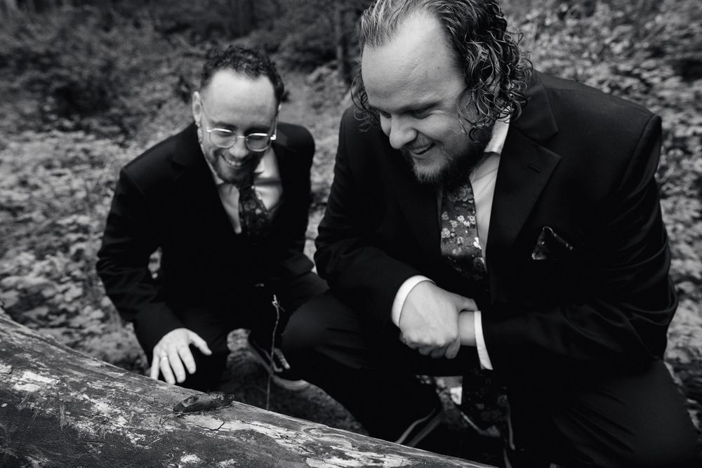 Two men in dark suits crouch outdoors in a black and white photo among rocks and trees.