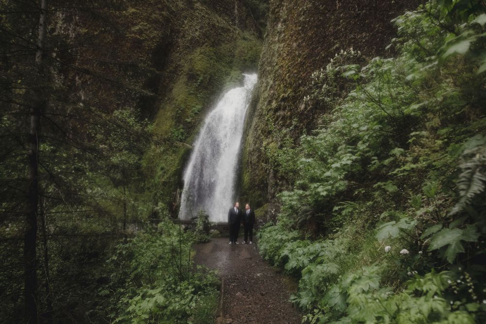 Two grooms stand on a forest trail before a cascading waterfall surrounded by lush green foliage. Wahkeena Falls in the Columbia River Gorge