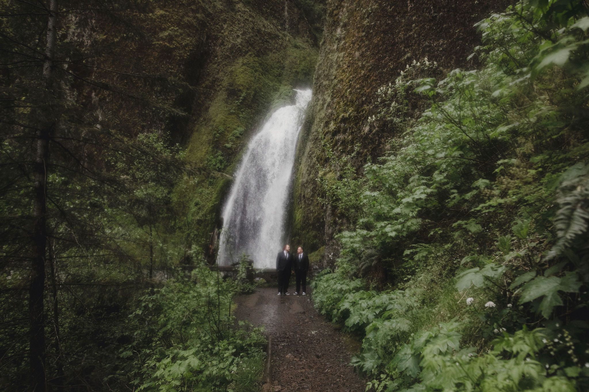 Two grooms stand on a forest trail before a cascading waterfall surrounded by lush green foliage. Wahkeena Falls in the Columbia River Gorge