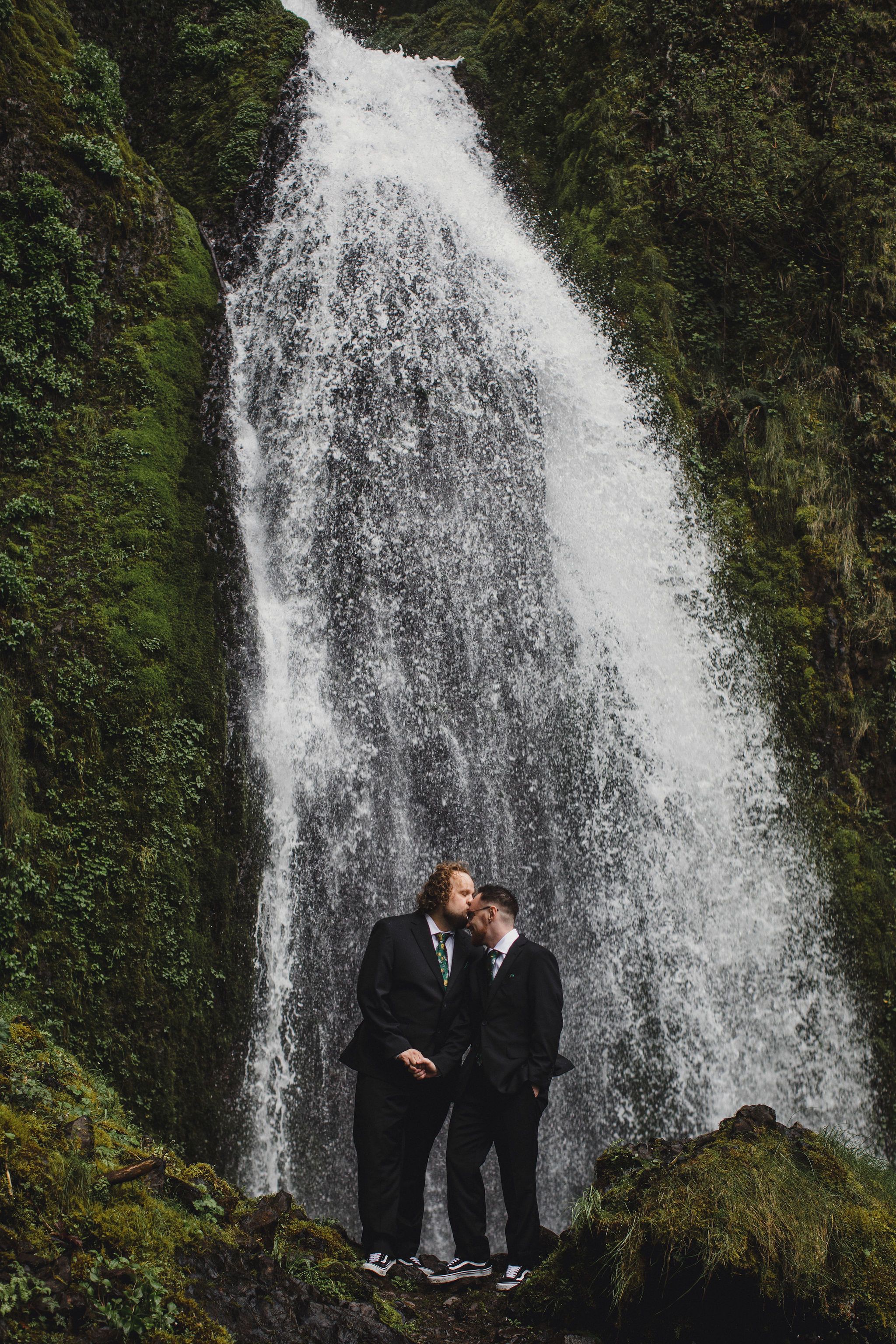 Two grooms in black suits stand romantically before a stunning waterfall surrounded by lush green moss. Columbia River Gorge, Wahkeena Falls