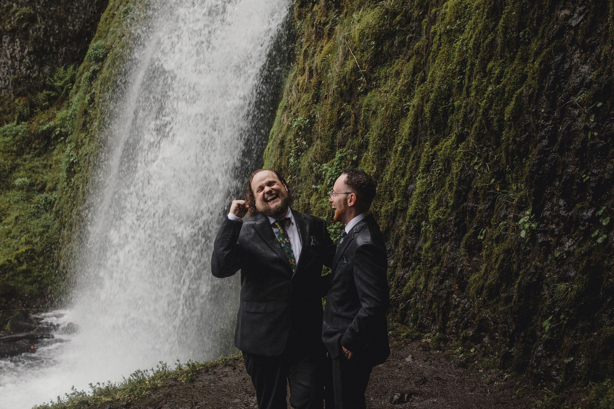 Two grooms laughing joyfully in front of a stunning mossy waterfall during their outdoor elopement at Wahkeena falls in oregon. .