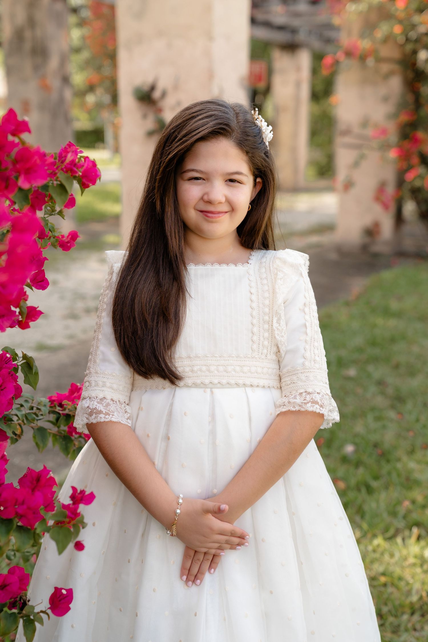 Young girl in white lace communion dress smiling outdoors near pink bougainvillea flowers.