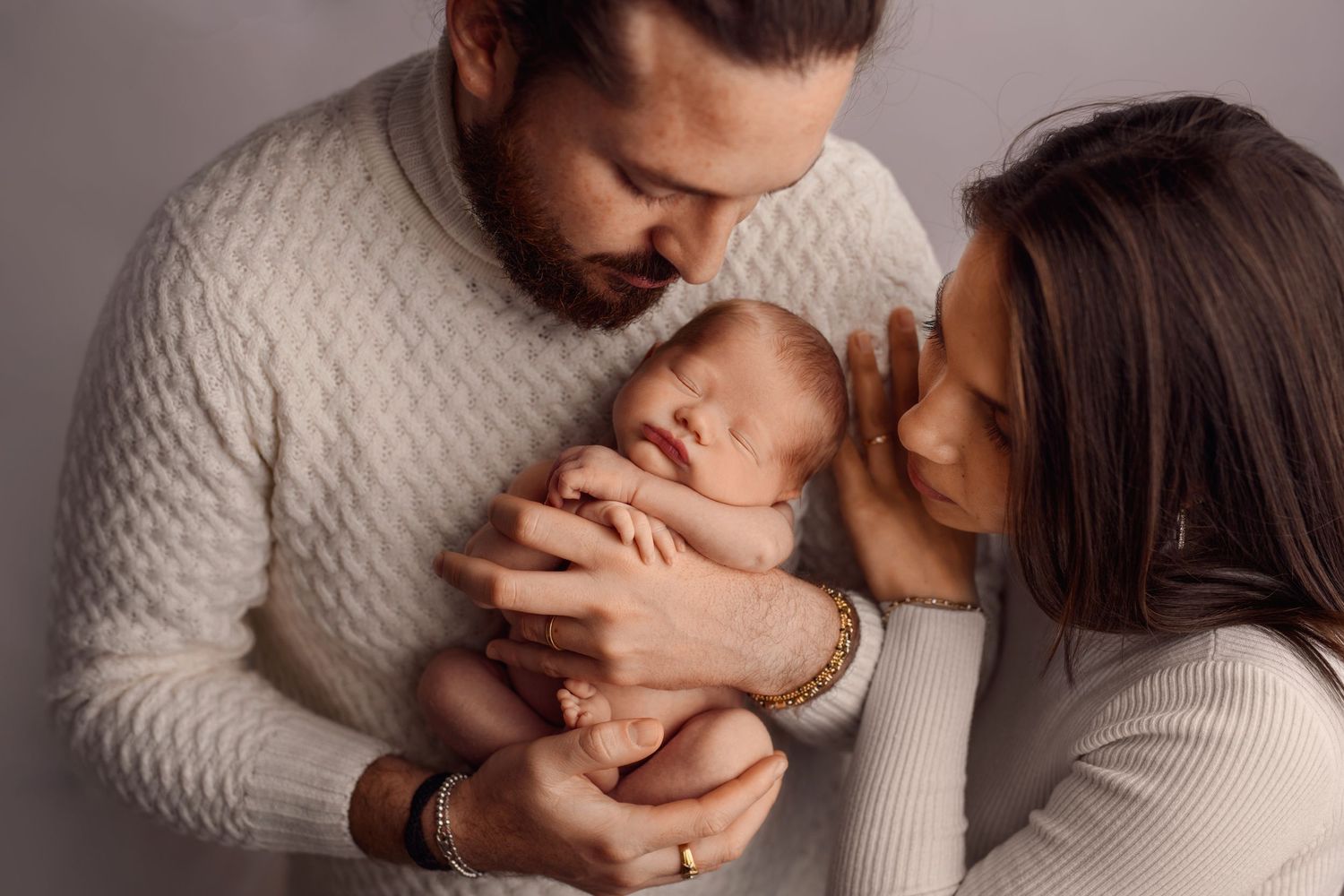 Parents in white outfits tenderly kiss their sleeping newborn baby during a studio portrait session.