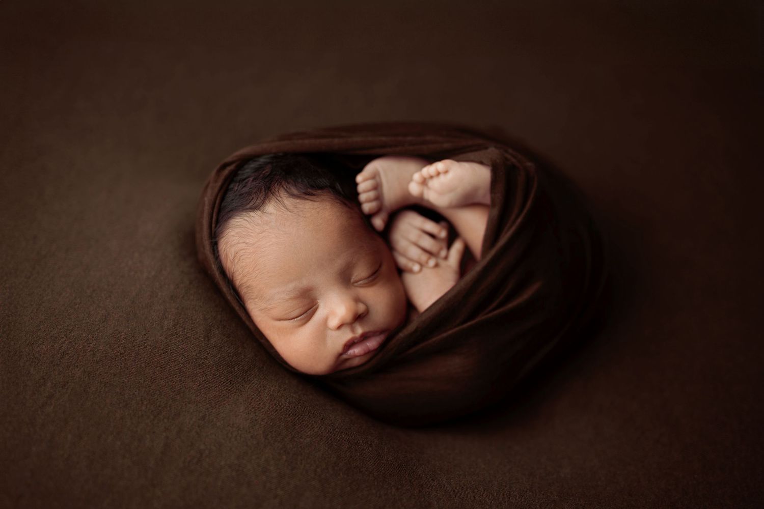 Newborn baby wrapped snugly in a brown swaddle blanket sleeping on a dark brown background.