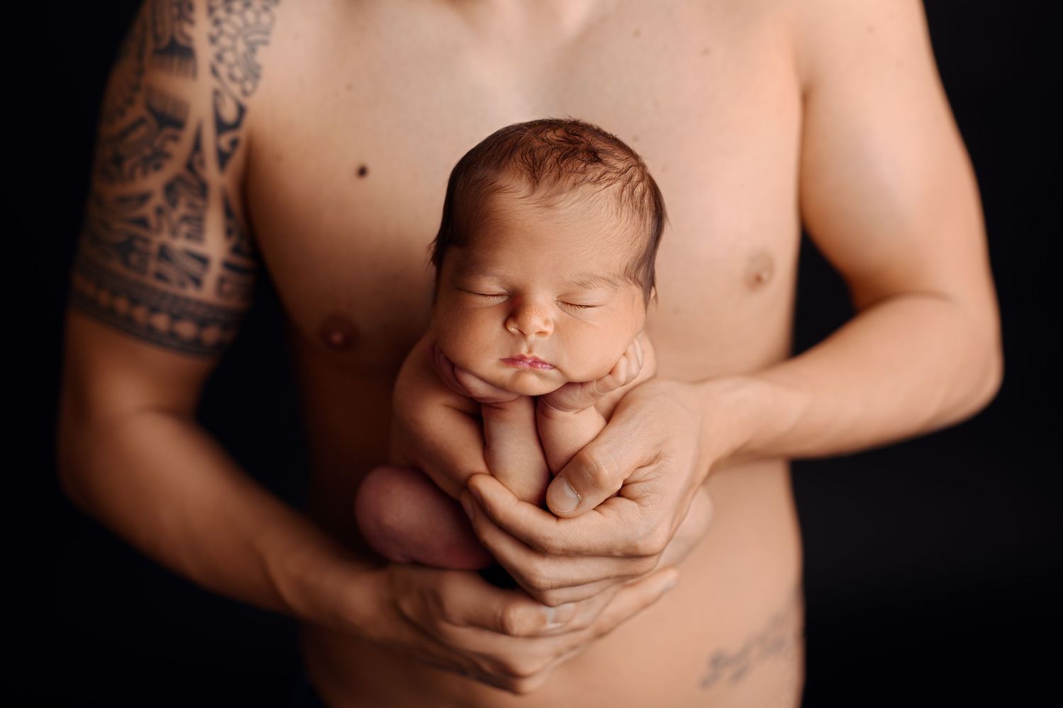 Tattooed father holds sleeping newborn baby gently in both hands against a black background.