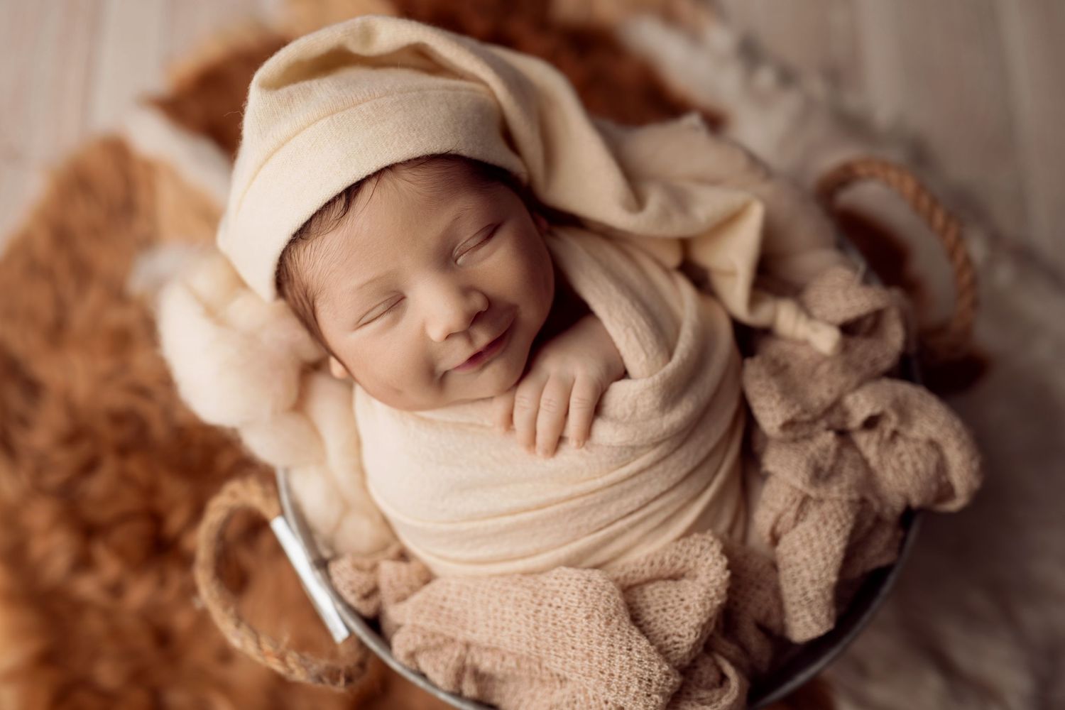 Smiling newborn baby wrapped in cream swaddle and knit hat sleeping peacefully in a metal bucket prop.