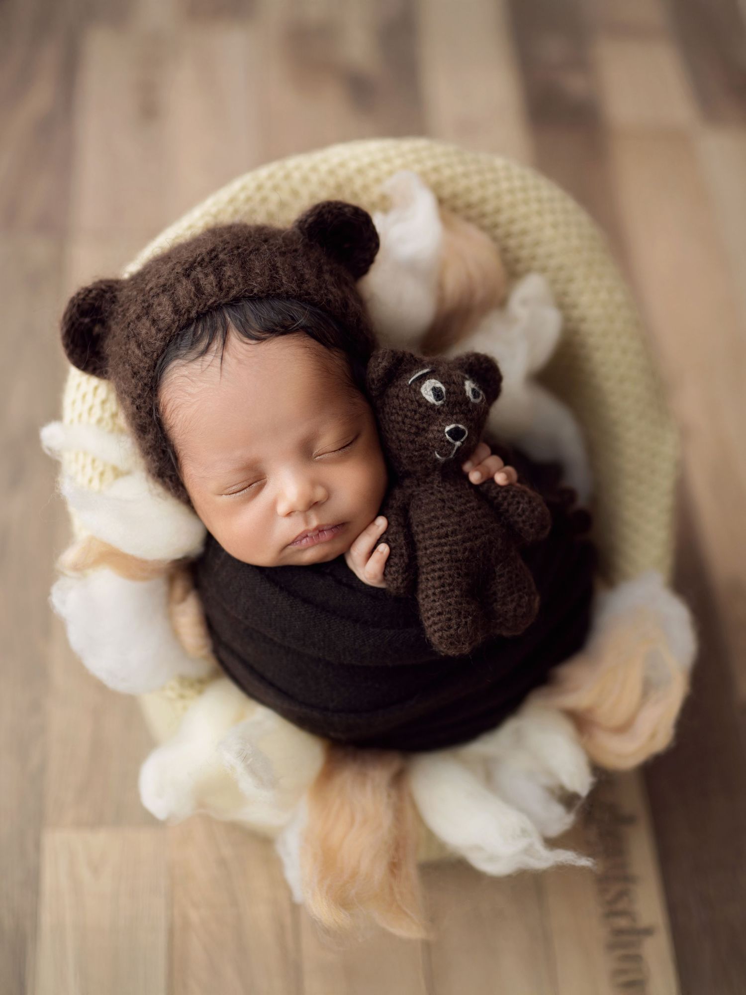 Newborn baby wearing a bear ear bonnet cuddles a knit teddy bear in a basket prop.