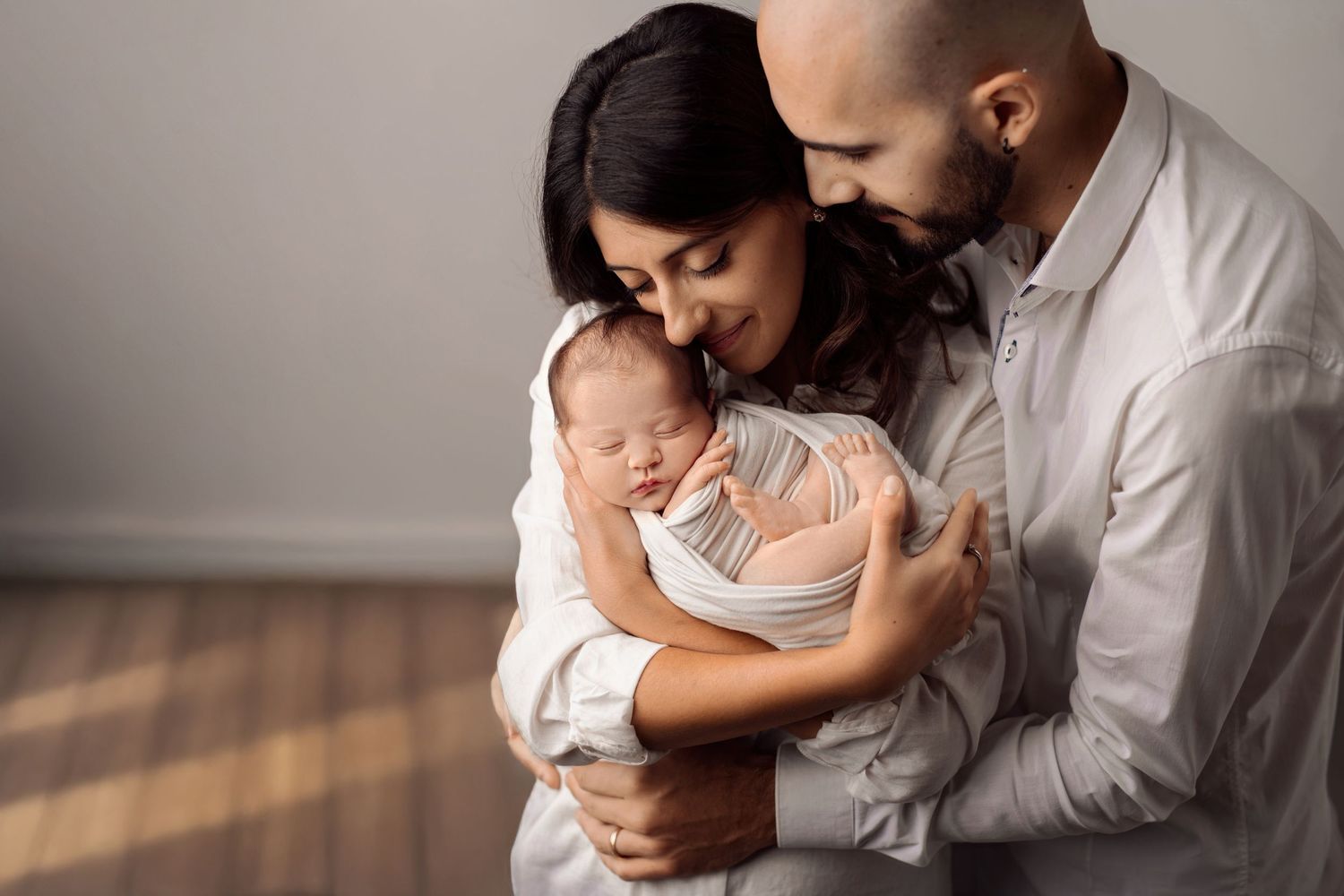 Loving couple in white clothing cuddle their swaddled newborn baby during an indoor portrait session.
