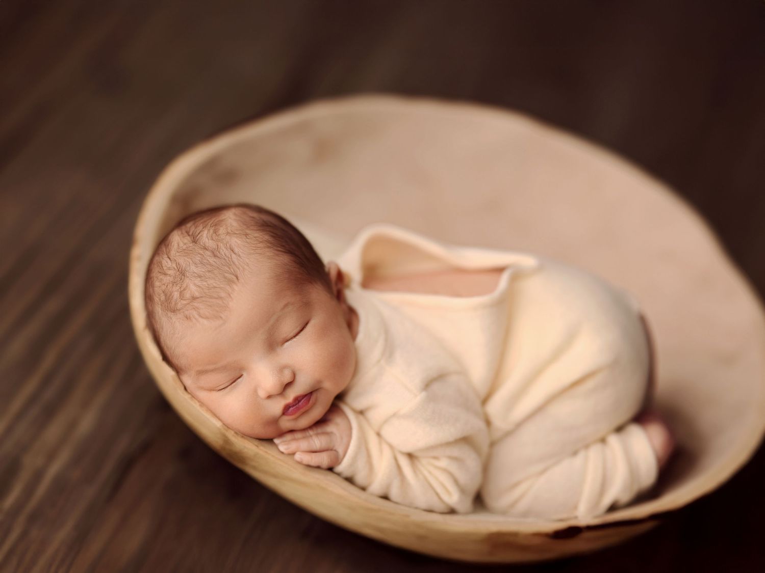 Peaceful newborn baby in cream outfit sleeps curled up inside a wooden bowl prop.