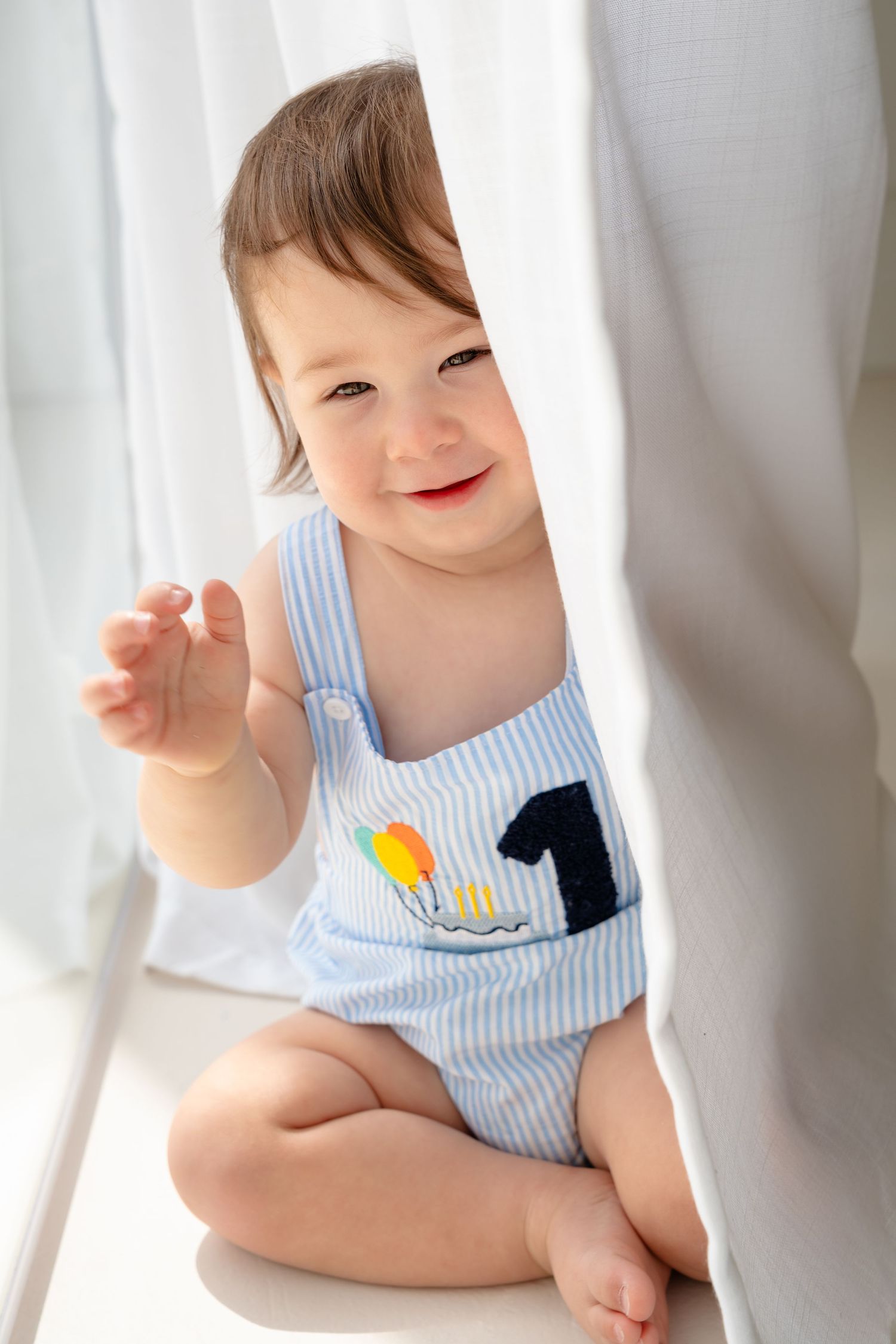Smiling baby in blue striped romper with number 1 applique sitting by white curtains in bright natural light.