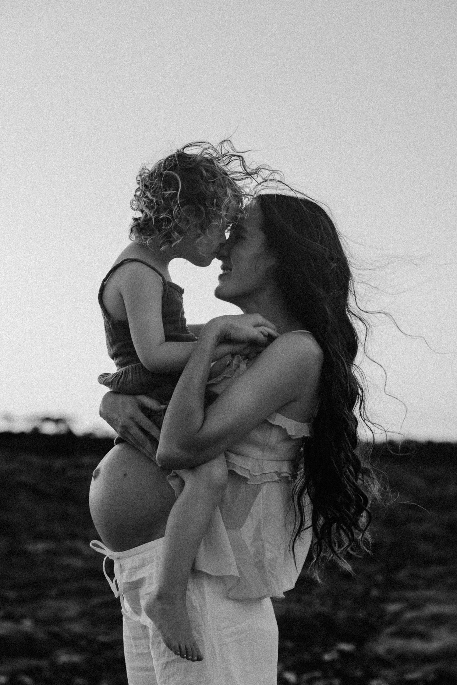 Black and white photo of a pregnant woman holding a curly-haired toddler, sharing a tender nose-to-nose moment outdoors.