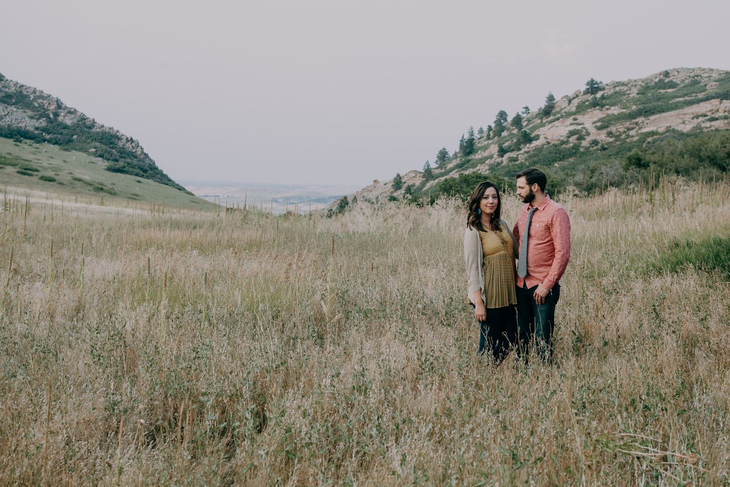 Couple standing together in a golden field with rolling hills in the background on an overcast day.