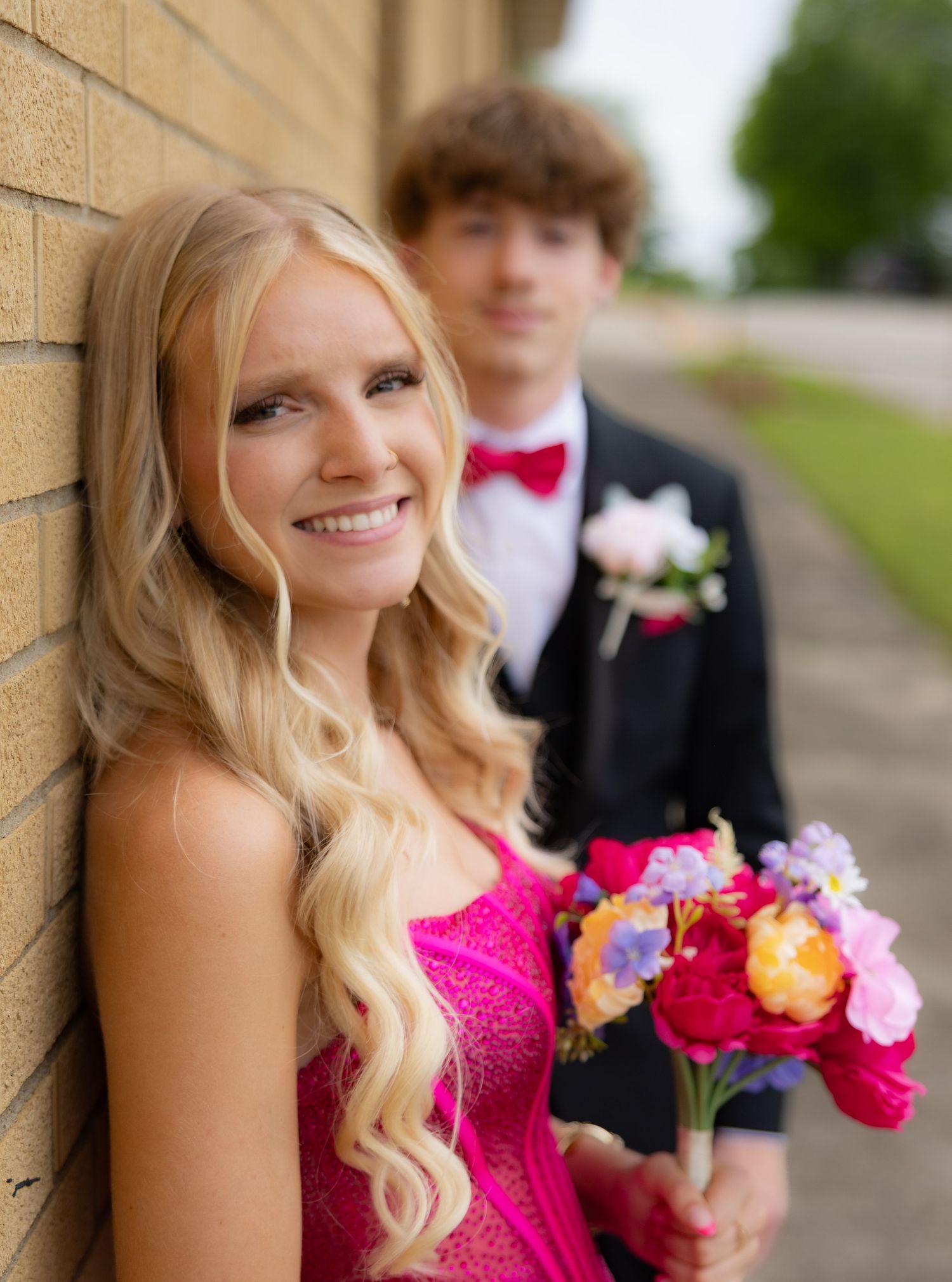 A smiling blonde teen in a pink prom dress holds colorful flowers beside a boy in a black tuxedo with red bow tie.