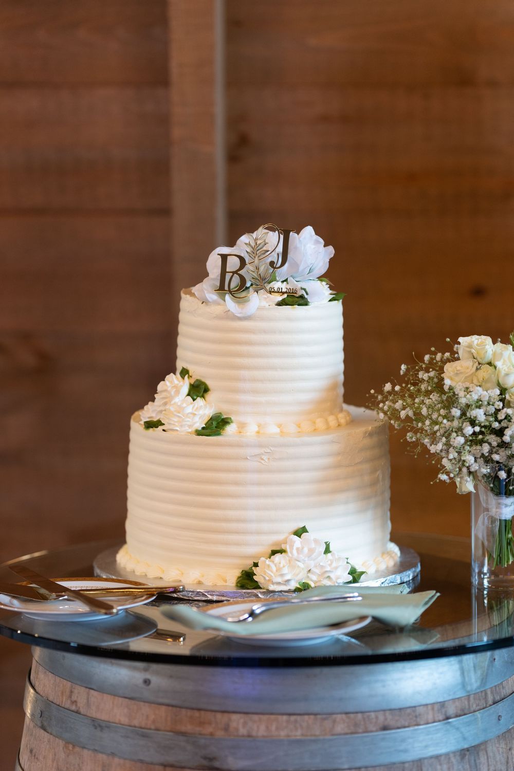 Two-tier white wedding cake with floral decorations and monogram topper on a barrel table.