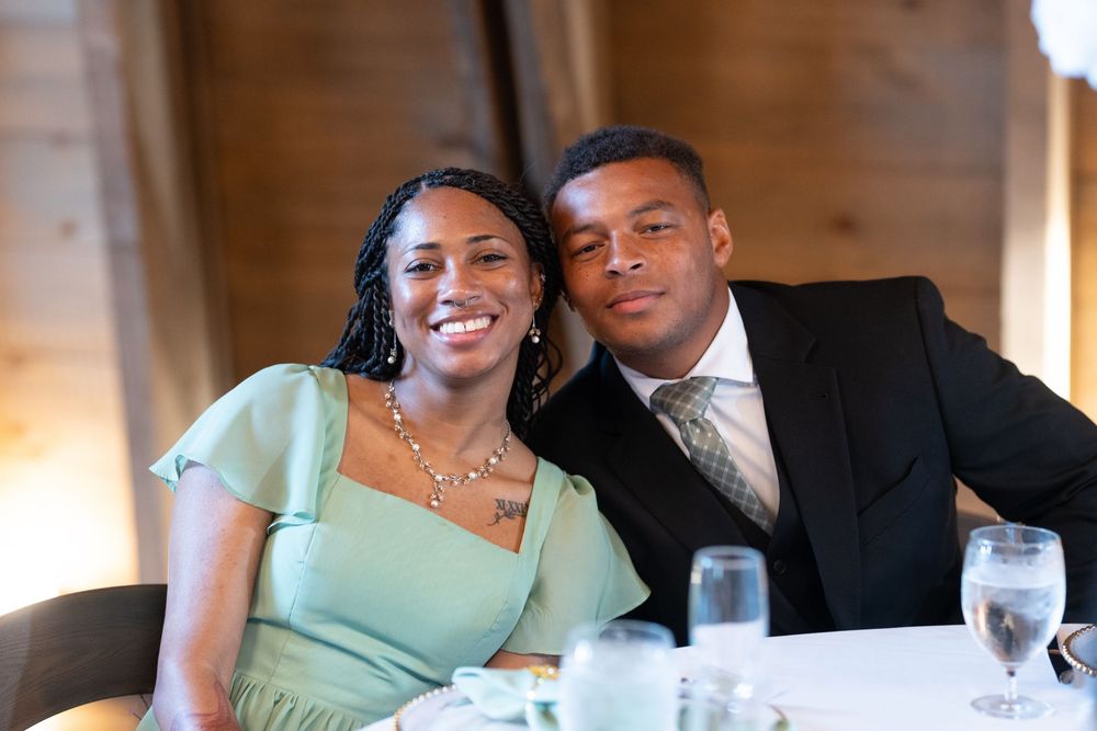 A smiling couple in formal attire poses together at a wedding reception table with water glasses nearby.