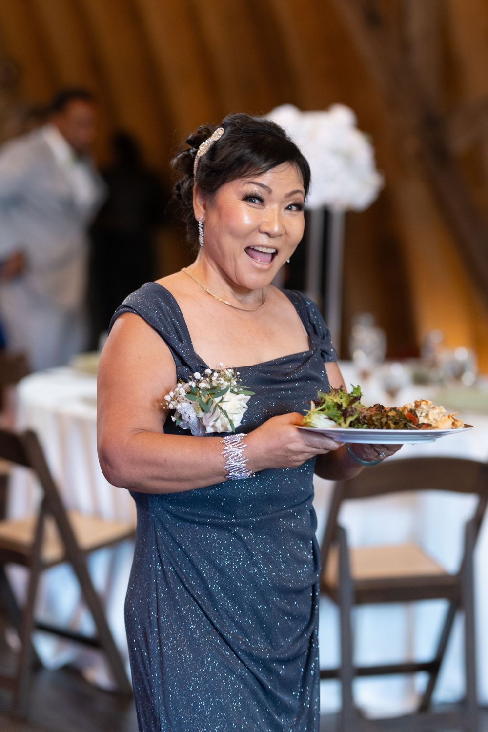 A woman in a sparkling gray gown with a wrist corsage carries a plate of food at a wedding reception.
