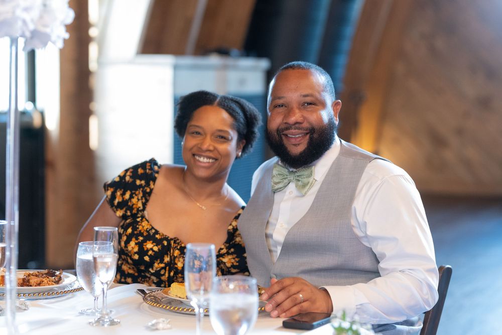 Smiling couple seated at a wedding reception table with glasses and elegant decor in a warmly lit venue.
