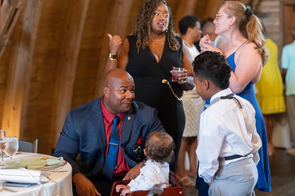 Adults and children socializing at an indoor wedding reception in a rustic barn venue.