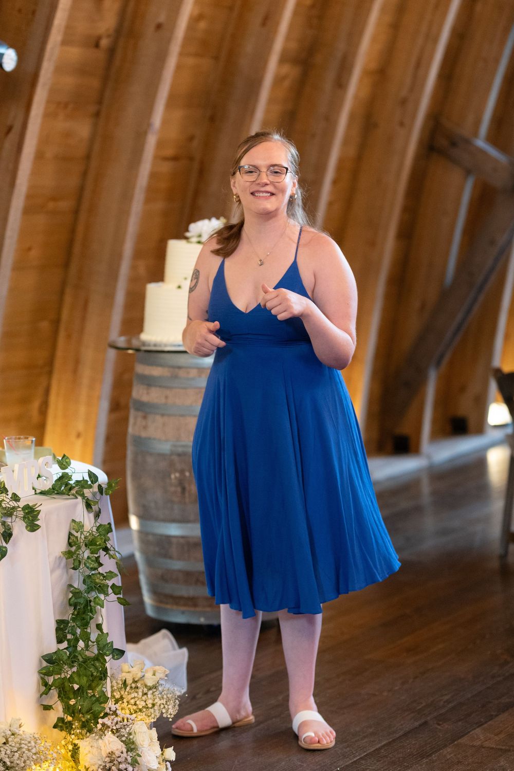 Smiling bridesmaid in blue midi dress poses near wedding cake in rustic barn venue.