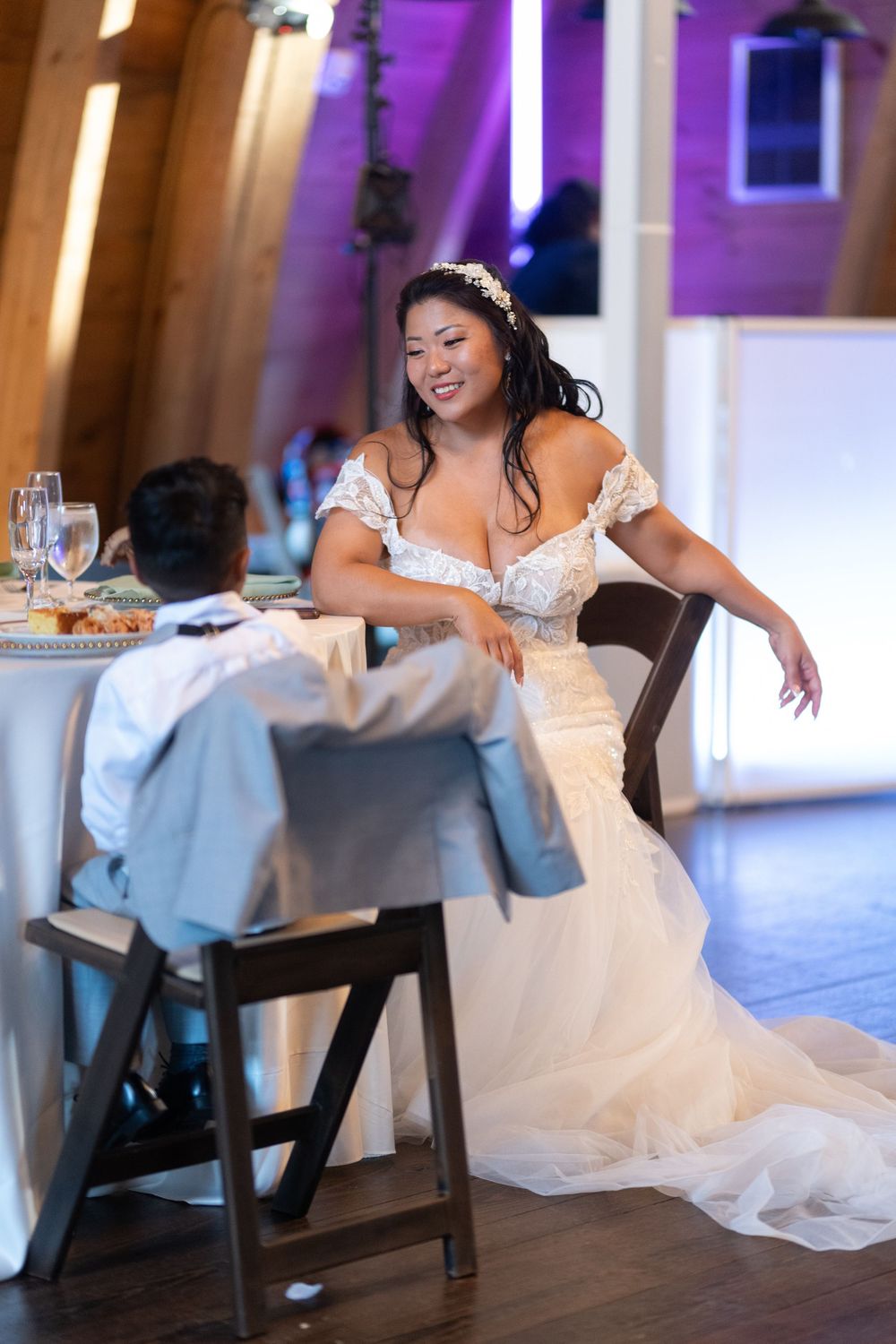 A bride in a white off-shoulder gown smiles at the groom during their wedding reception dinner.