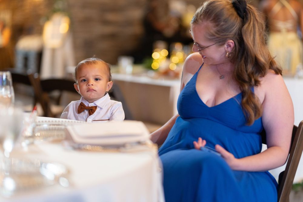 A baby in a bow tie sits at a wedding reception table beside a pregnant woman in a blue dress.