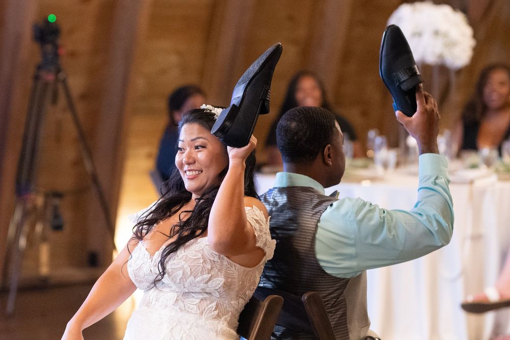 Bride and groom laughing while holding up shoes during fun wedding reception game at rustic barn venue.