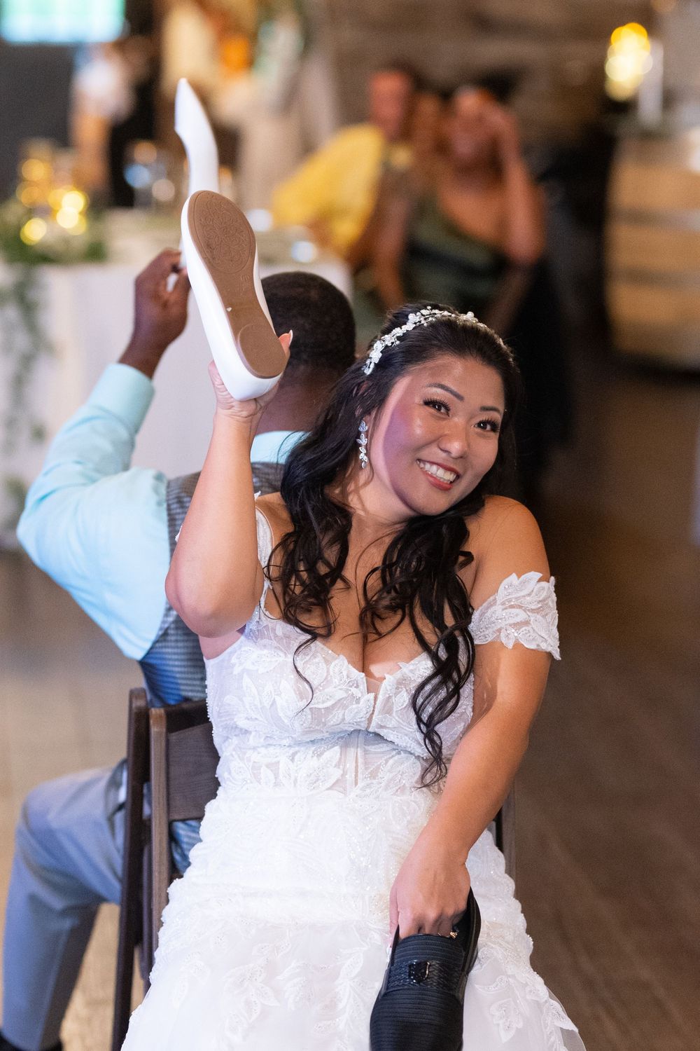 Bride in white lace gown holds up groom's shoe during wedding reception shoe game at Sweeney Barn.