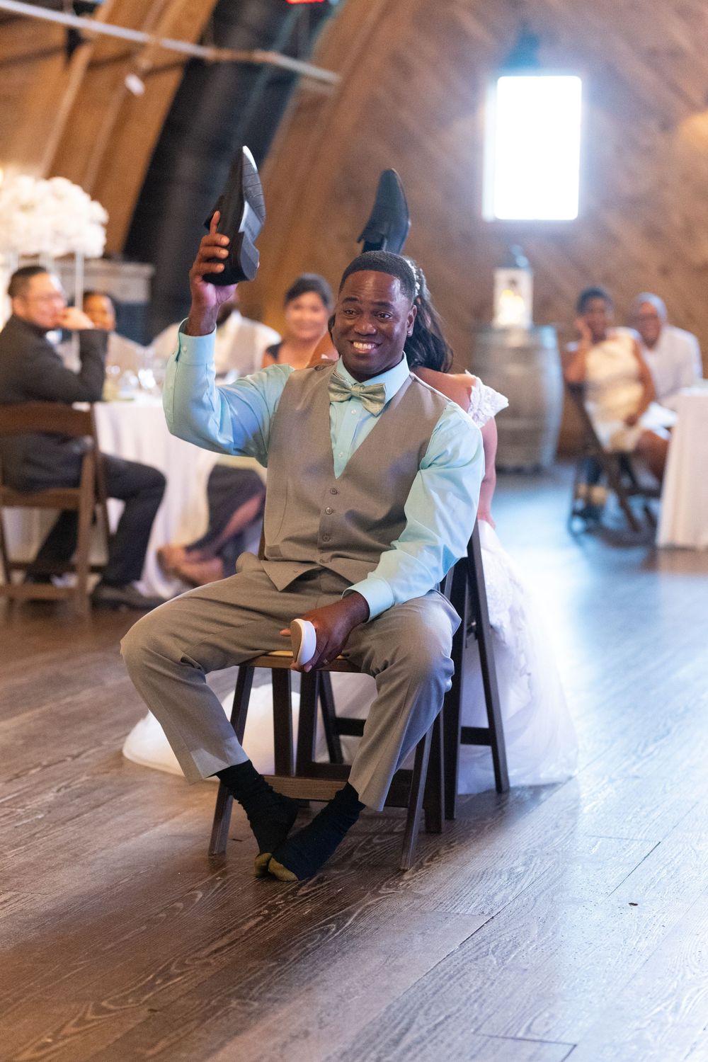 Groom holds up shoe during wedding reception shoe game while seated in chair at rustic barn venue.