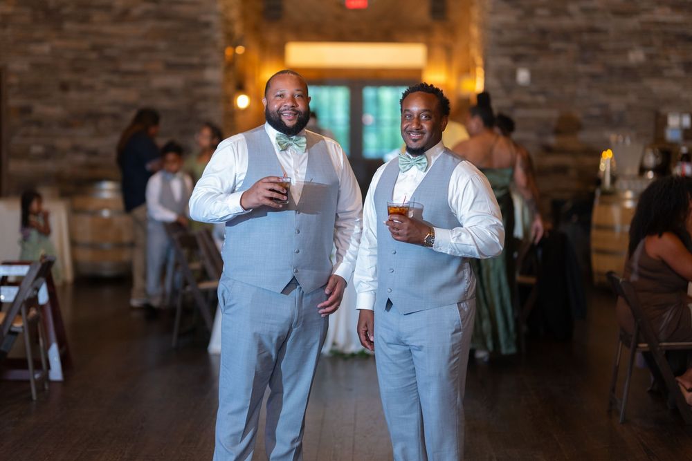 Two groomsmen in light blue suits with bow ties smile holding drinks at an elegant wedding reception venue.
