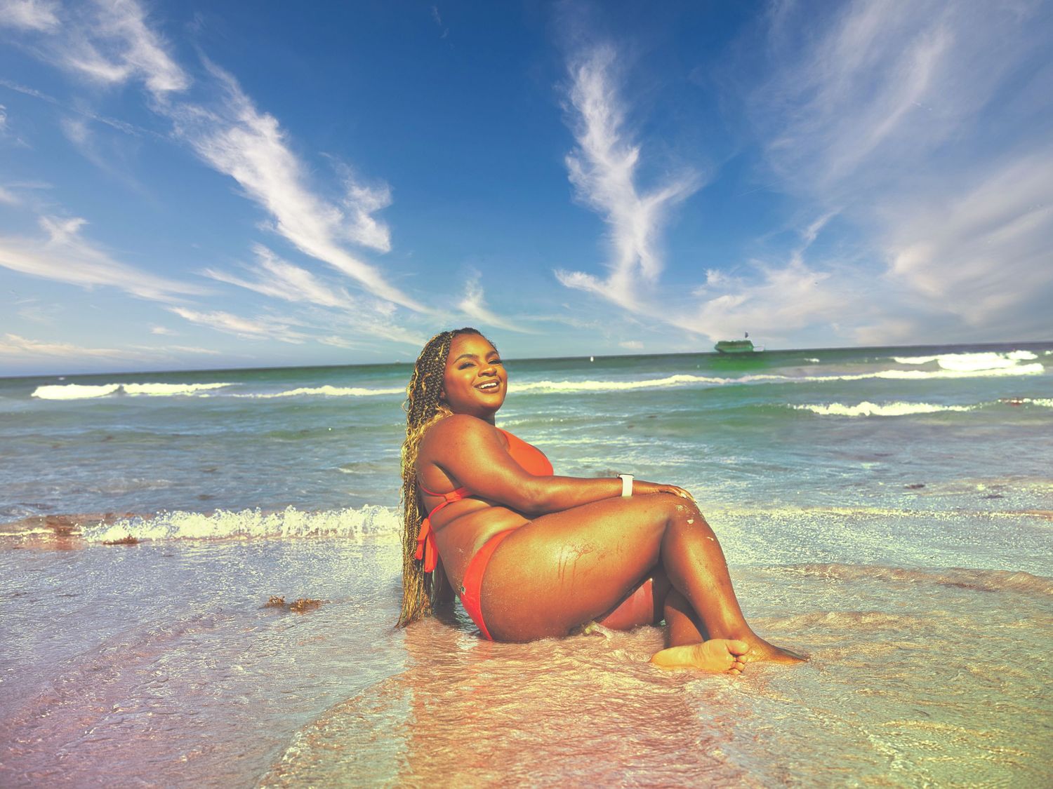 Woman in red bikini sitting in shallow ocean waves on a tropical beach under a vibrant blue sky.