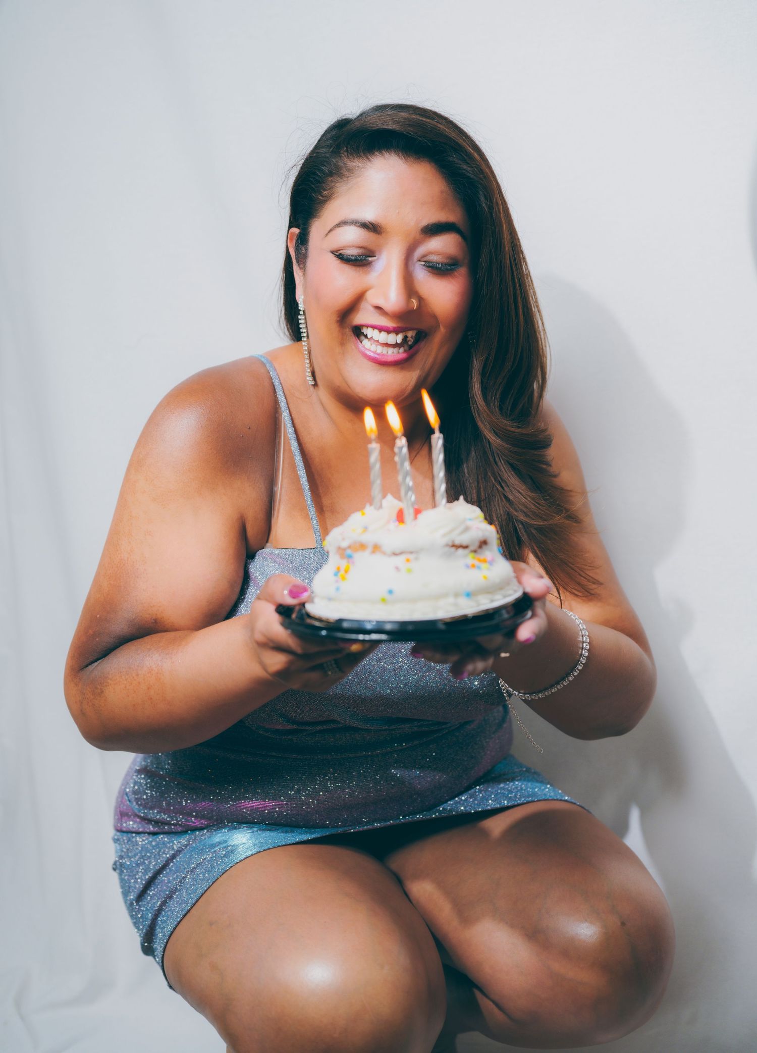 Smiling woman in sparkly dress holding a small birthday cake with lit candles, celebrating her special day.