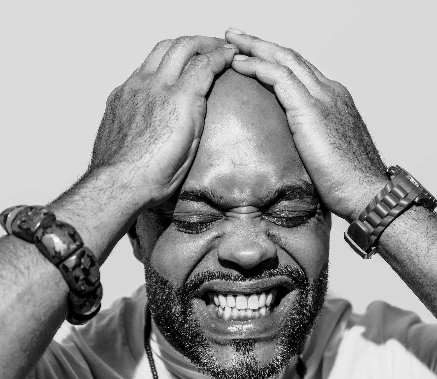 Black and white photo of a man laughing with eyes closed, hands on head, wearing bracelets.