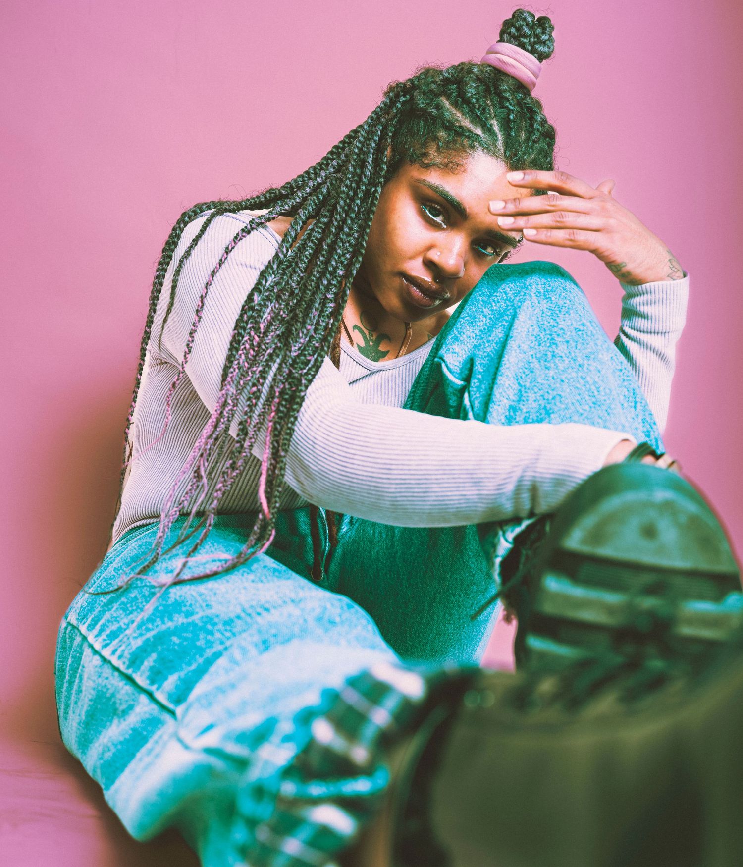 Young Black woman with long braids poses against pink background wearing blue overalls and white top.