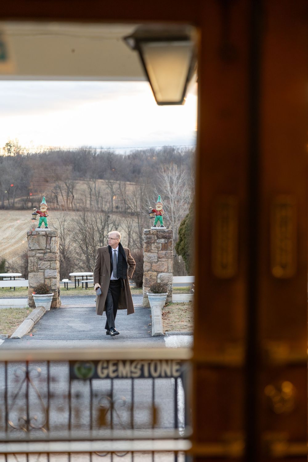 Elderly man in long coat walks toward stone gate pillars with elf statues at a Gemstone estate.
