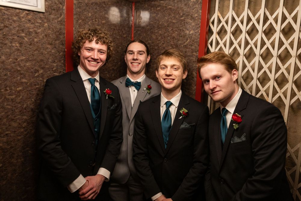 Four young men in formal suits with boutonnieres smiling together at a formal event or prom.
