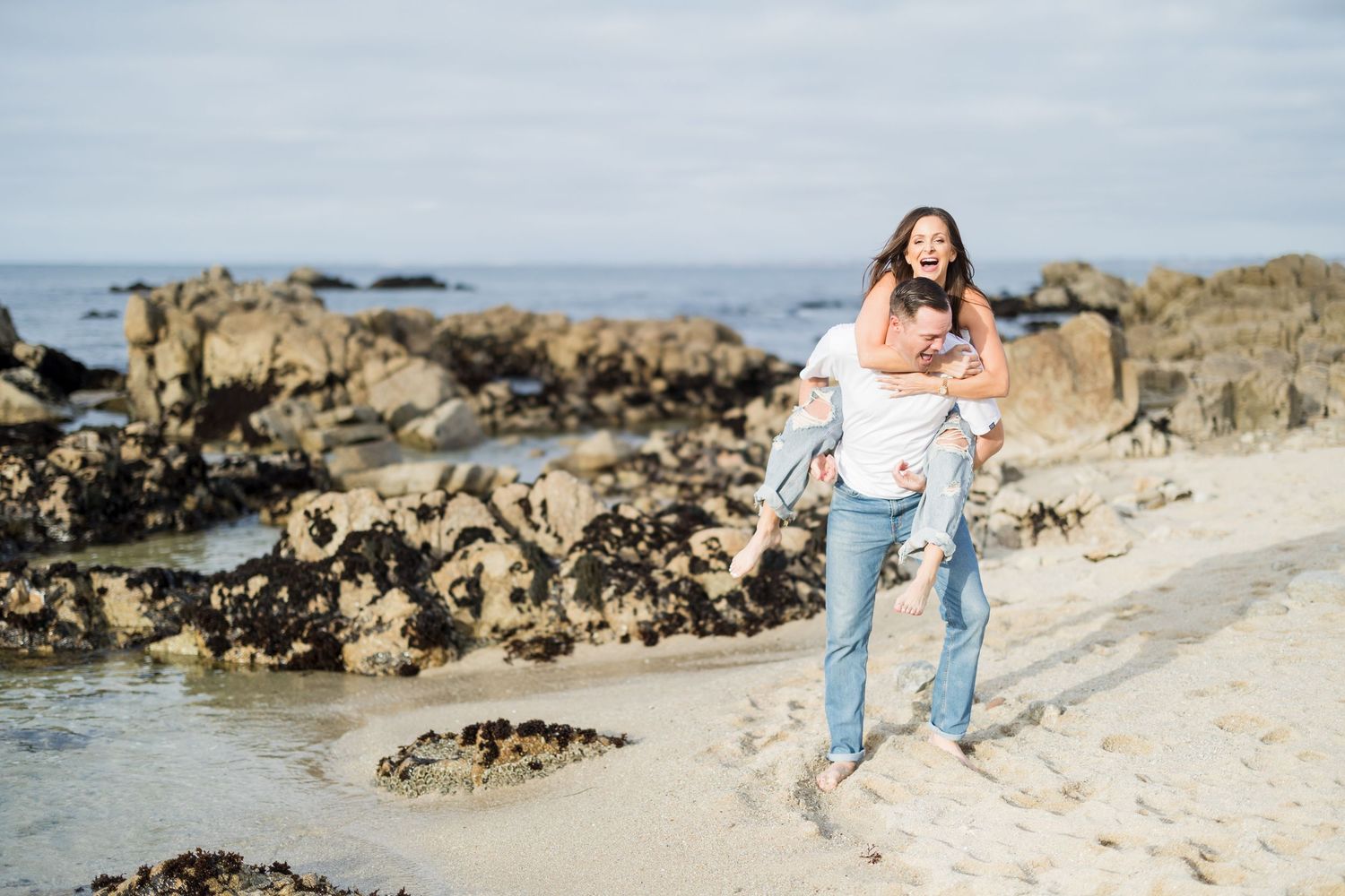 Couple embracing on a scenic hilltop with ocean and mountains visible in golden sunlight behind them.
