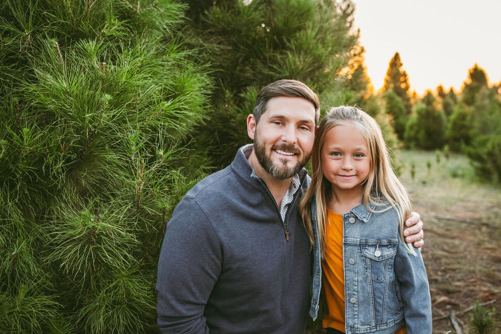 A smiling man and young girl pose together in front of lush green pine trees at a Christmas tree farm.