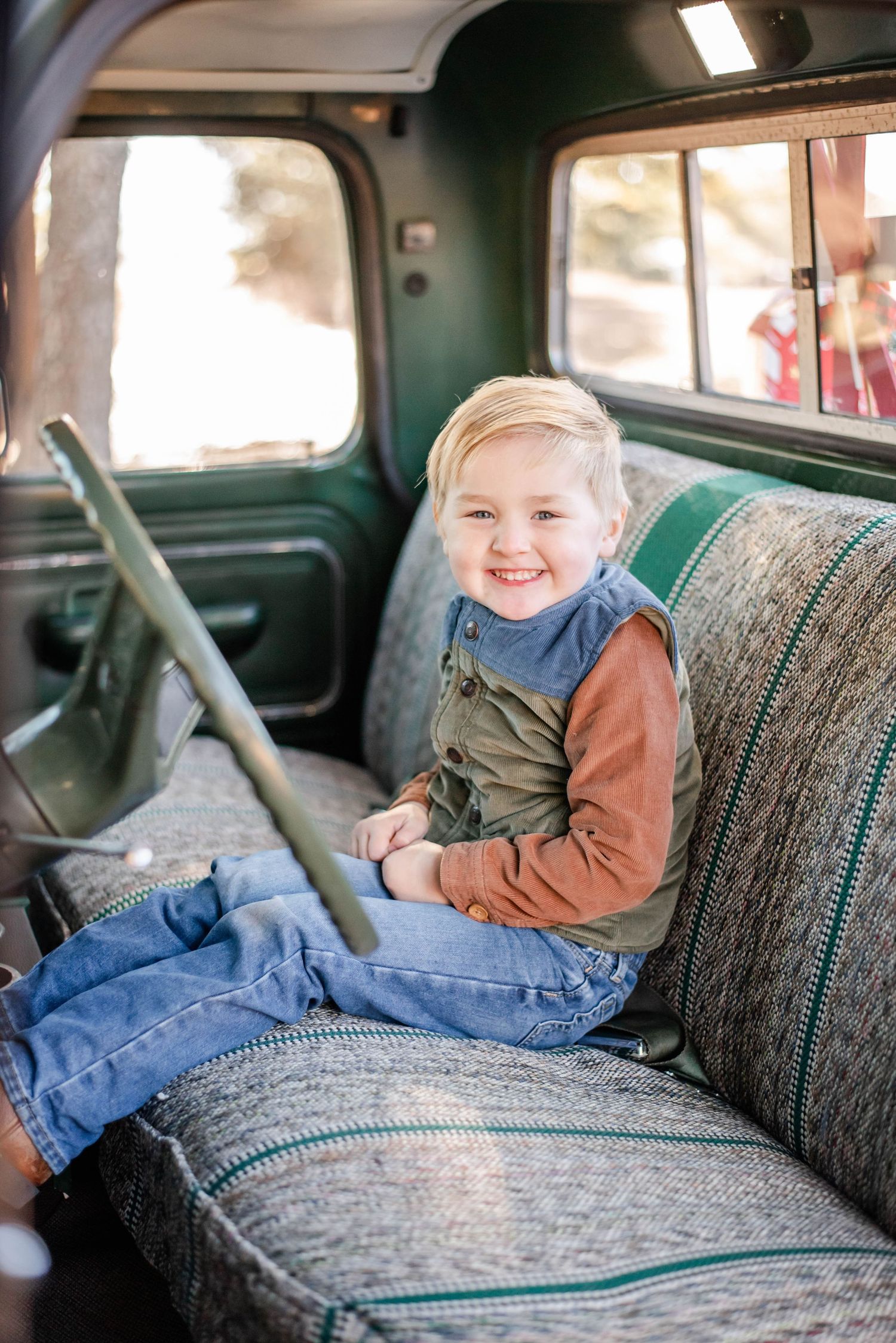 Smiling blonde toddler boy sitting on a gray bench seat inside a vintage green truck cab.