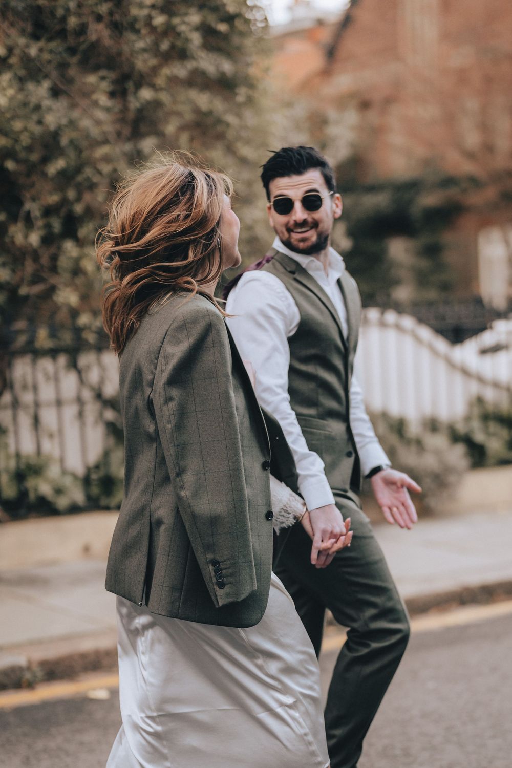 A stylish couple walks hand in hand on a street, the man in a vest and sunglasses smiling back at the camera.