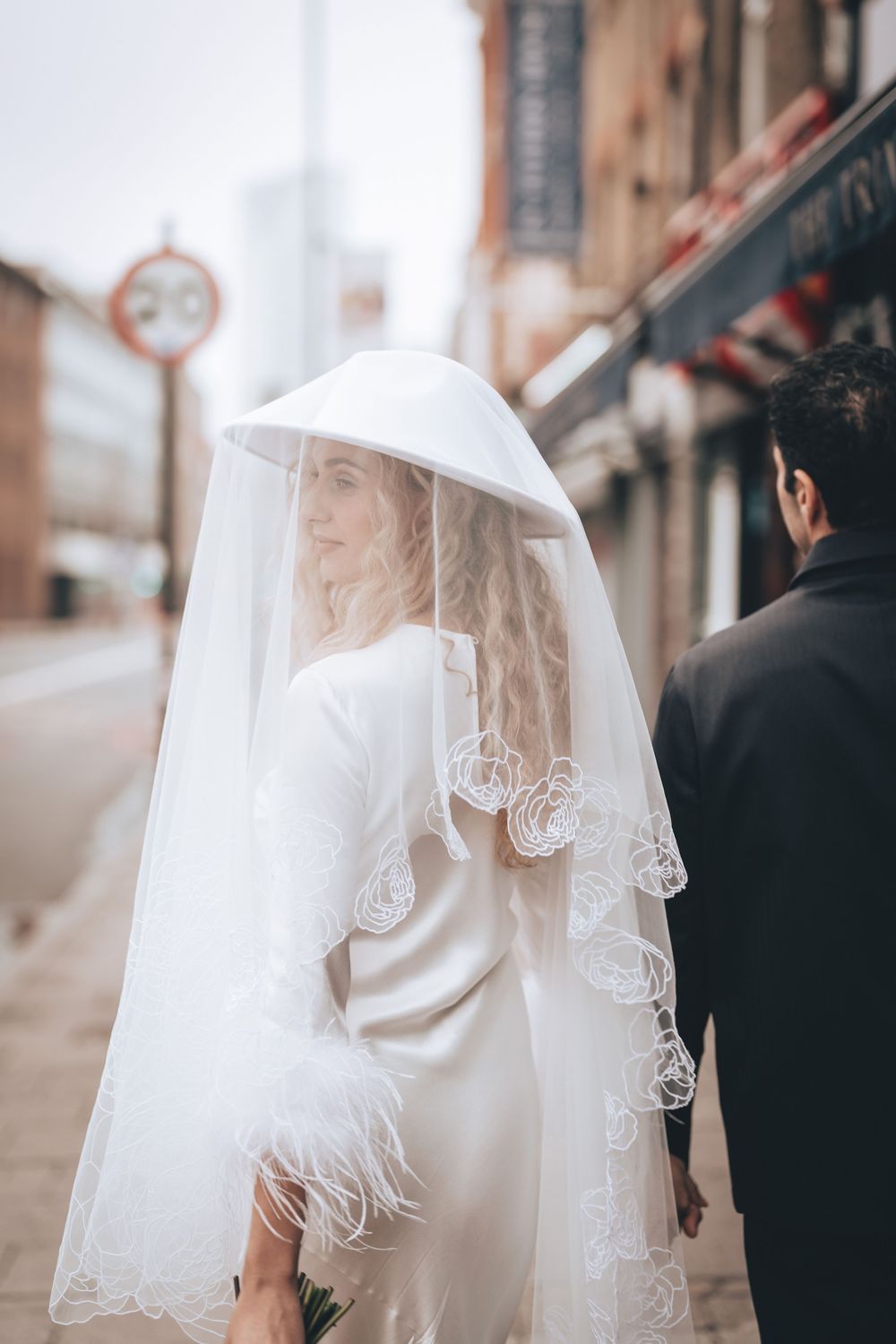 Bride in white satin gown with feather trim and lace veil walks beside groom in black suit on city street.