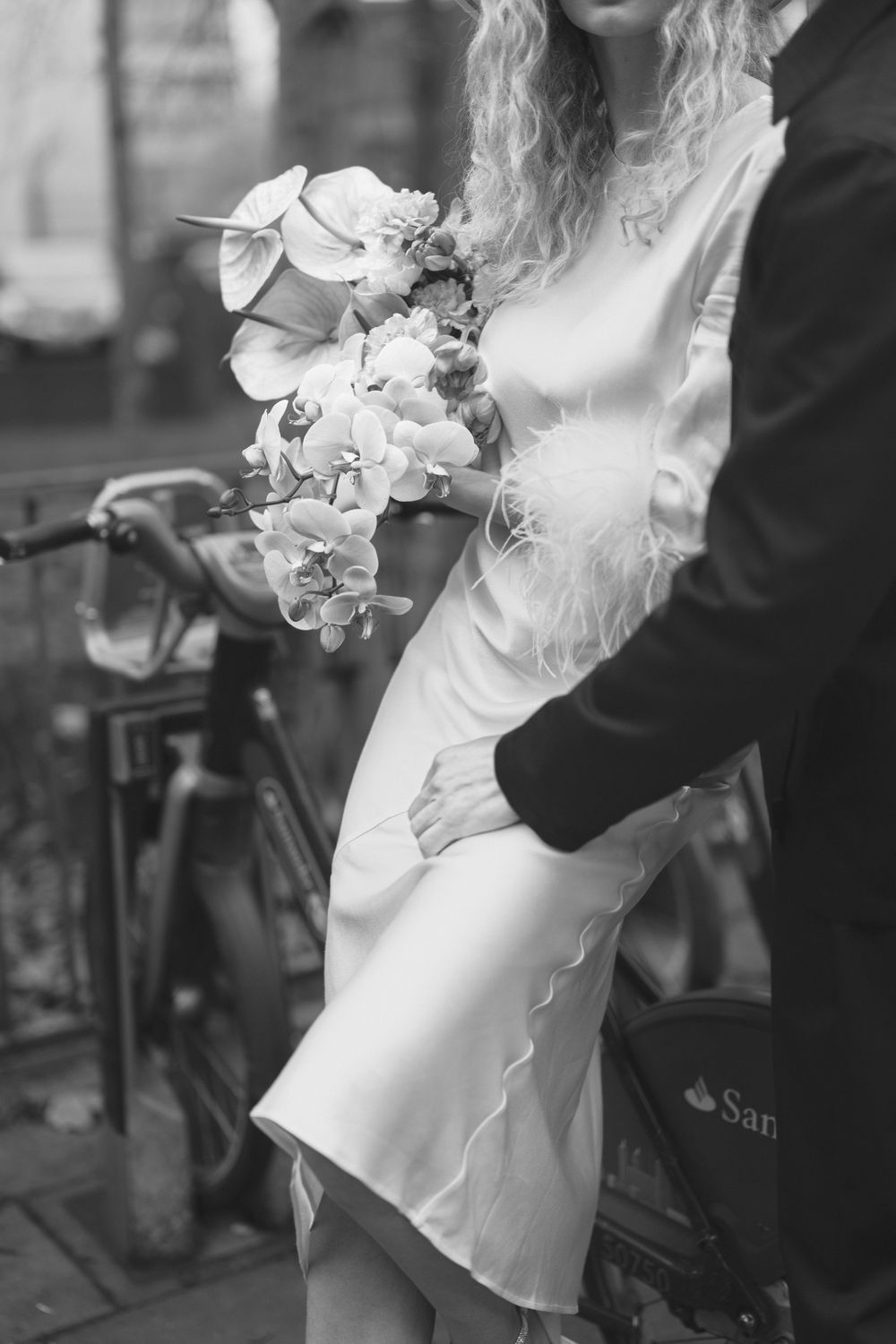 Black and white photo of a bride in a satin dress holding a floral bouquet, embraced by groom near bicycles.