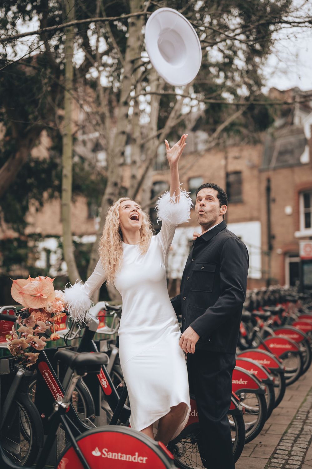 Bride in white dress tosses hat near Santander bikes while groom watches, London street wedding photo. London elopement wedding.