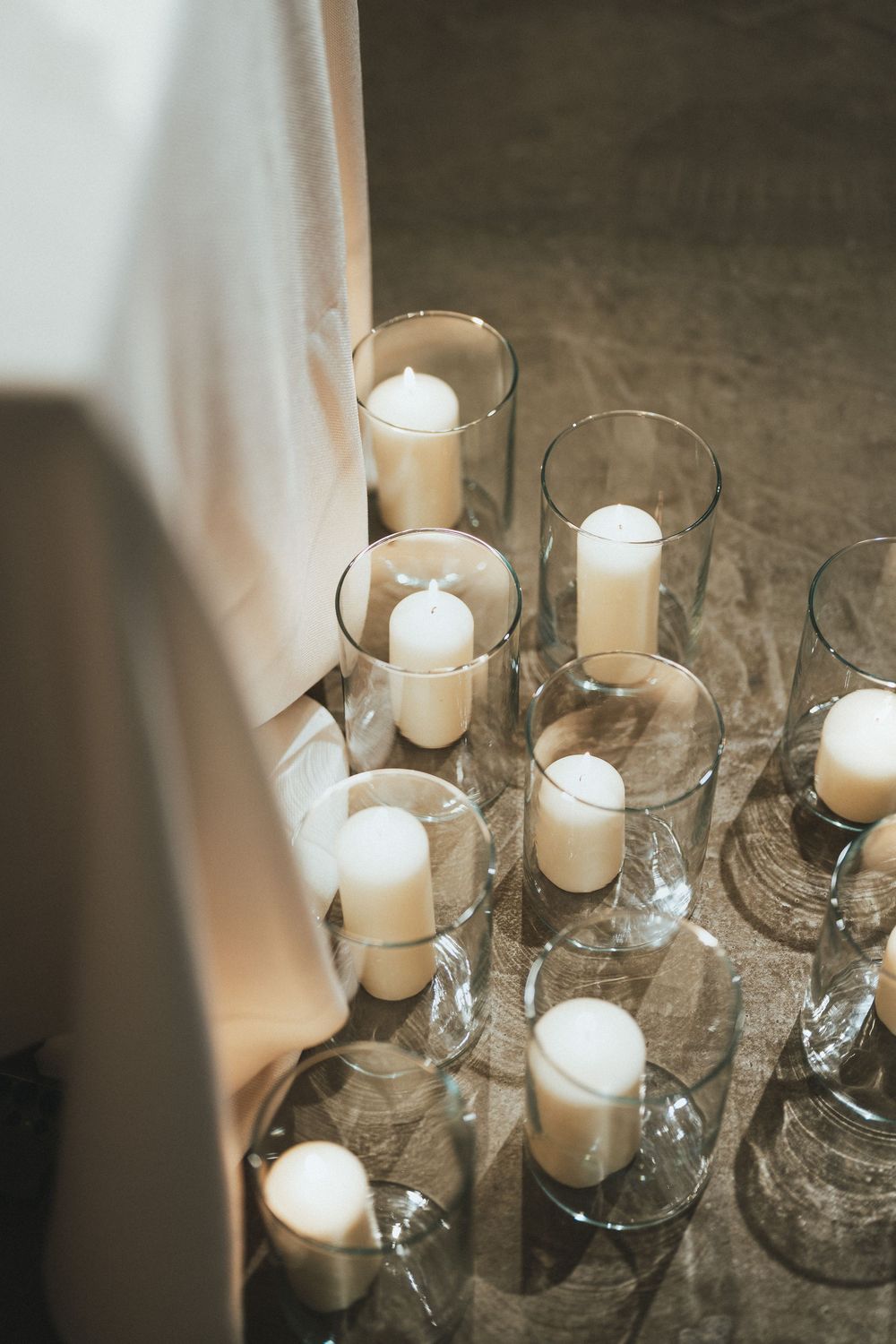 White pillar candles in glass hurricane vases arranged on floor beside draped fabric at elegant event.