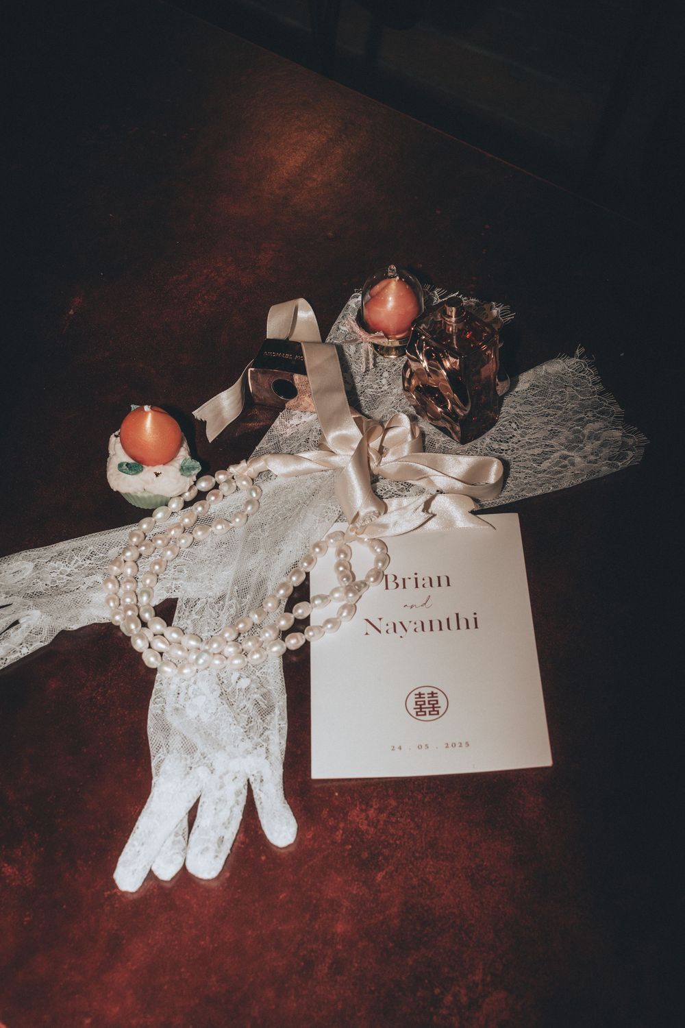 Wedding flat lay with lace gloves, pearl necklace, candles, and Brian & Nayanthi ceremony program on dark surface.