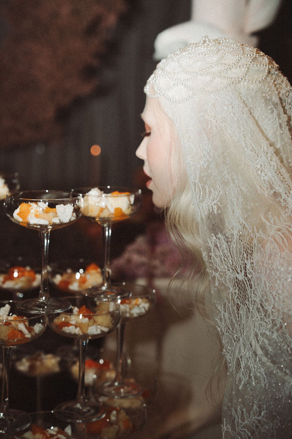 Bride in white lace veil admiring elegant champagne tower with fruit desserts at wedding reception. Vintage Film 35 photograph.