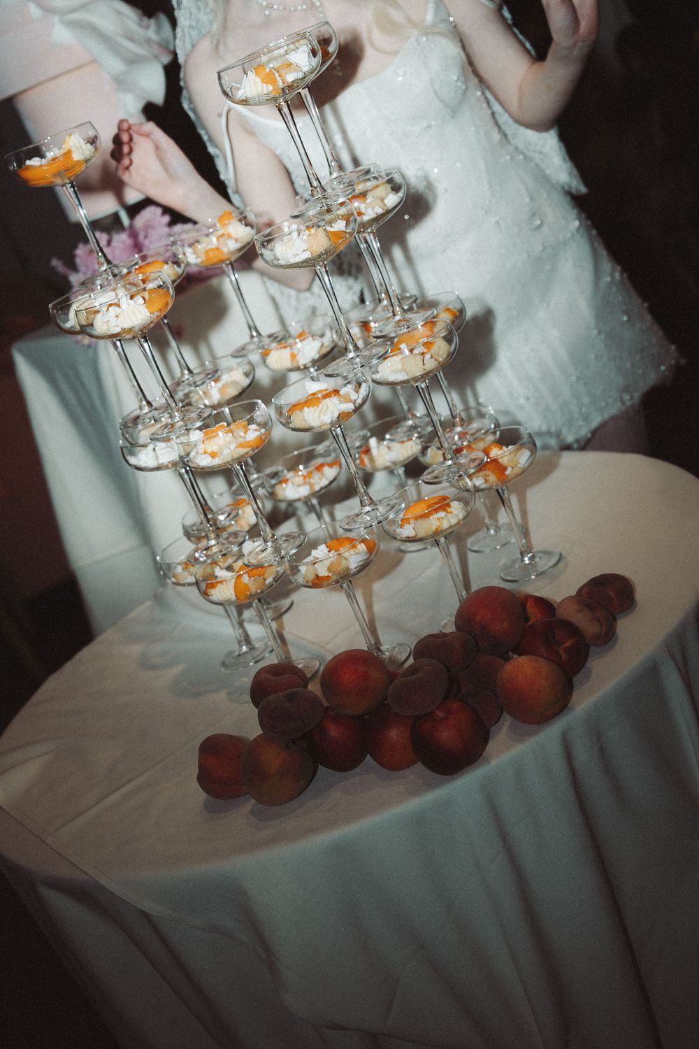 Elegant champagne tower with peach desserts on stemmed glasses, surrounded by fresh peaches on a white tablecloth.
