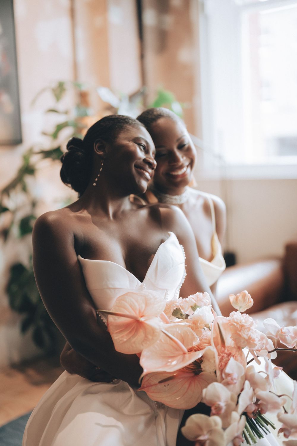 Two Black brides in white gowns smile joyfully, holding a bouquet of blush pink anthuriums and orchids indoors.